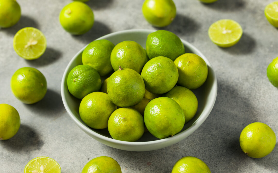 Fresh green limes in a bowl on a gray background, with whole and halved limes scattered around.