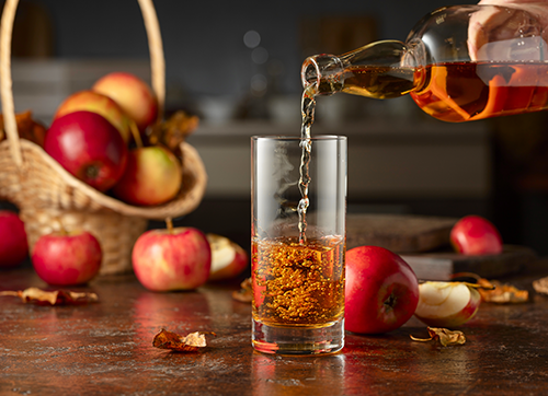 Apple juice being poured from a bottle into a glass. There is a basket full of apples in the background.