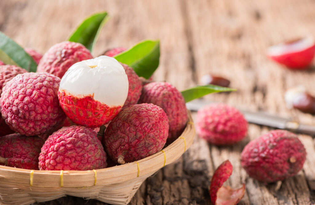 Basket of fresh lychees with one peeled lychee revealing its juicy white flesh, placed on a rustic wooden surface, showcasing the vibrant fruit used for Lychee Puree.